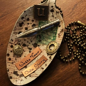 Op de afbeelding: Een wit keramisch gerecht in de vorm van een blad met een bruin, zwart en gouden ontwerp. Het gerecht heeft een metalen penpunt, een gouden knoop en twee koperen labels met de tekst "Write Your Own Story". Er ligt een gouden ketting naast het gerecht.