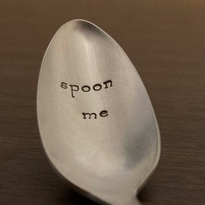 May include: A silver spoon with the words "spoon me" stamped into the bowl. The spoon is made of a shiny metal and has a long handle. The spoon is on a dark brown surface.