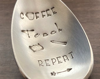 May include: A silver spoon with the words "Coffee Teach Repeat" engraved on it. The spoon also has a small coffee cup, a book, a pencil, and an arrow engraved on it.