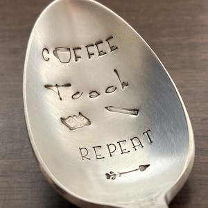 May include: A silver spoon with the words "Coffee Teach Repeat" engraved on it. The spoon also has a small coffee cup, a book, a pencil, and an arrow engraved on it.