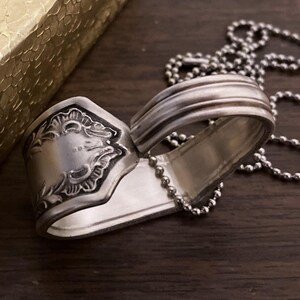 May include: A silver spoon necklace with a heart-shaped design. The spoon has a decorative pattern on the handle and is attached to a silver chain.