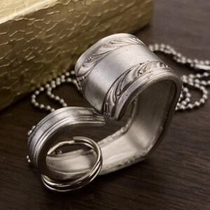 May include: A silver heart-shaped pendant necklace made from a repurposed spoon. The pendant has a detailed floral design and is attached to a silver ball chain.