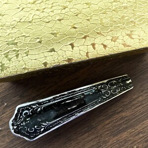 May include: A gold box with a textured surface and a black metal spoon with an ornate design.