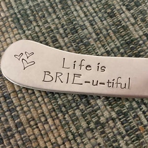 May include: Silver cheese spreader with the phrase "Life is BRIE-u-tiful" engraved on the blade, along with two small heart illustrations. The spreader has a decorative handle and is designed for serving cheese.