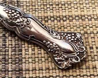 May include: A silver key chain made from a vintage silverware handle with a grapevine design. The handle is attached to a silver key ring.