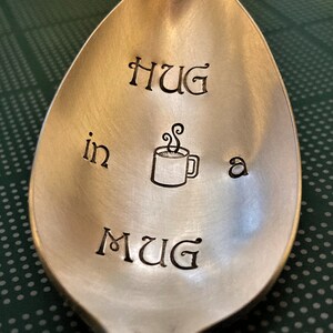 May include: A silver spoon with the words "HUG in a MUG" engraved on it. The spoon also has a small drawing of a mug with steam coming out of it.