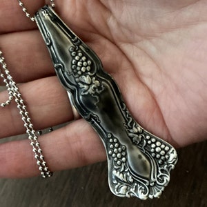 May include: A silver necklace with a vintage spoon pendant featuring a grapevine design. The spoon is oxidized and has a dark patina.