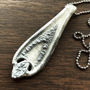 May include: A silver spoon pendant necklace with an ornate design. The spoon is attached to a silver ball chain.