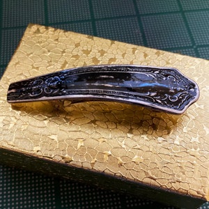 May include: A silver-toned hair clip with an ornate design. The clip is black and has a decorative pattern. The clip is sitting on a gold-colored surface.