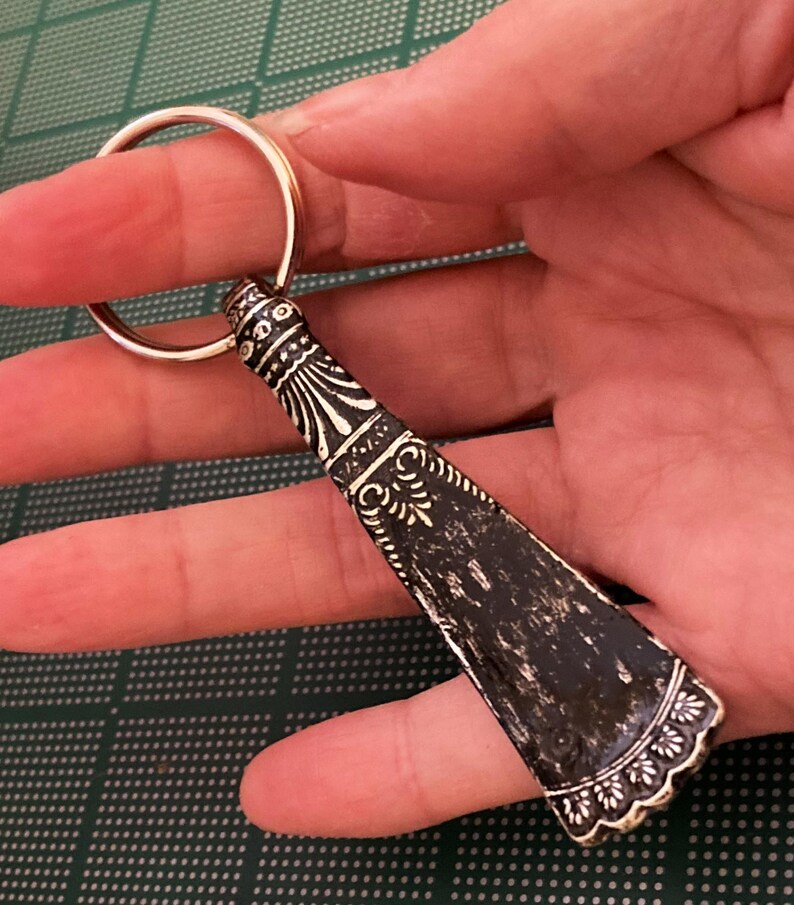May include: A silver key chain with a decorative black and white design. The key chain is shaped like a small shoe horn.