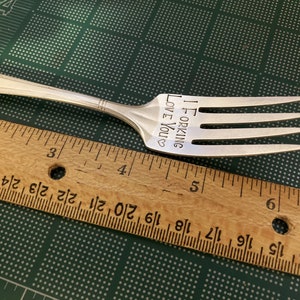 May include: A silver fork with the words "I FORKING LOVE YOU" engraved on the handle. The fork is lying on a green surface with a wooden ruler next to it.