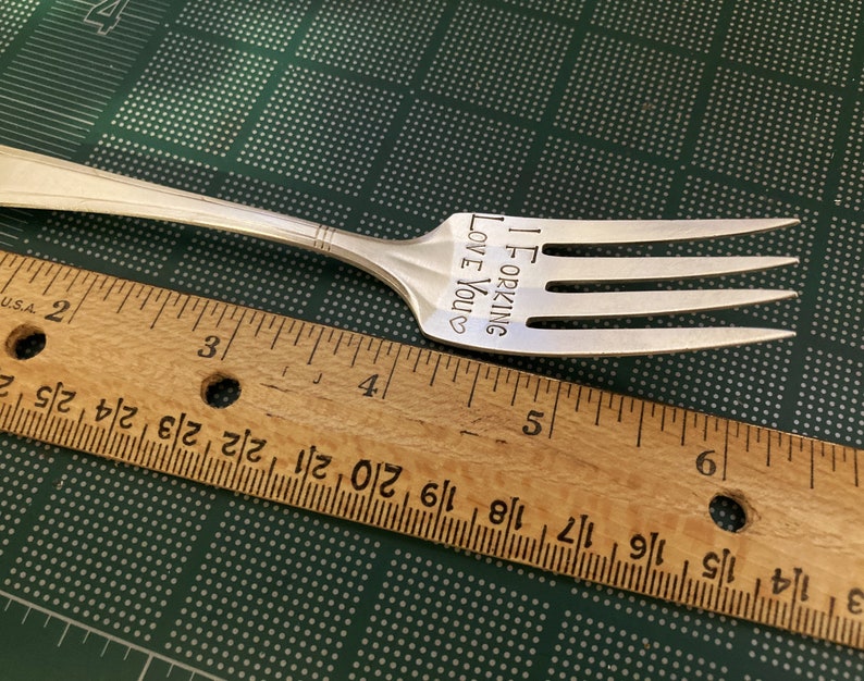 May include: A silver fork with the words "I FORKING LOVE YOU" engraved on the handle. The fork is lying on a green surface with a wooden ruler next to it.