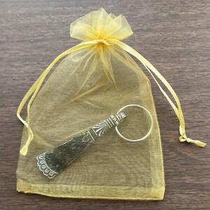 May include: A silver shoe horn with an ornate design is displayed in a yellow organza bag with a drawstring closure.