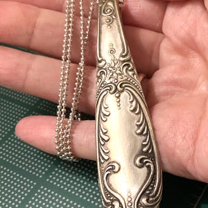 May include: A silver spoon necklace with an ornate design. The spoon is attached to a silver chain.