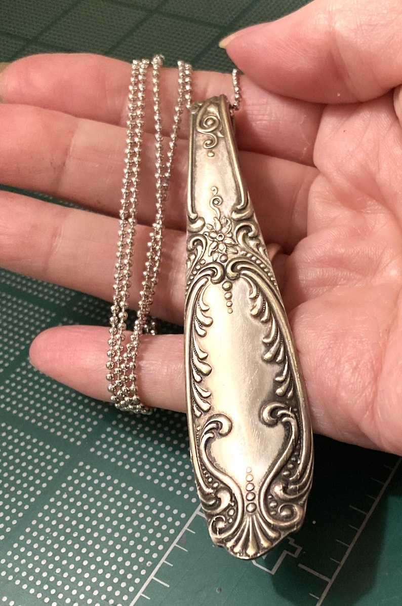 May include: A silver spoon necklace with an ornate design. The spoon is attached to a silver chain.