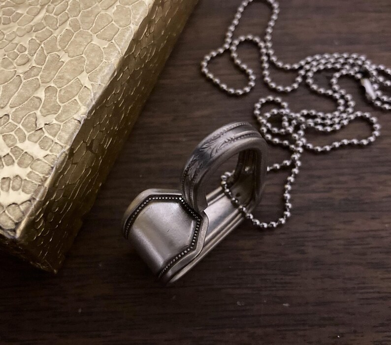 May include: A silver spoon necklace with a decorative design. The spoon is bent into a heart shape and is attached to a silver ball chain.
