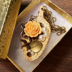 May include: A delicate necklace with a cream-colored pendant featuring a peach-colored rose, a bird, and the word "BALANCE". The pendant is surrounded by pearls and is attached to a gold chain.