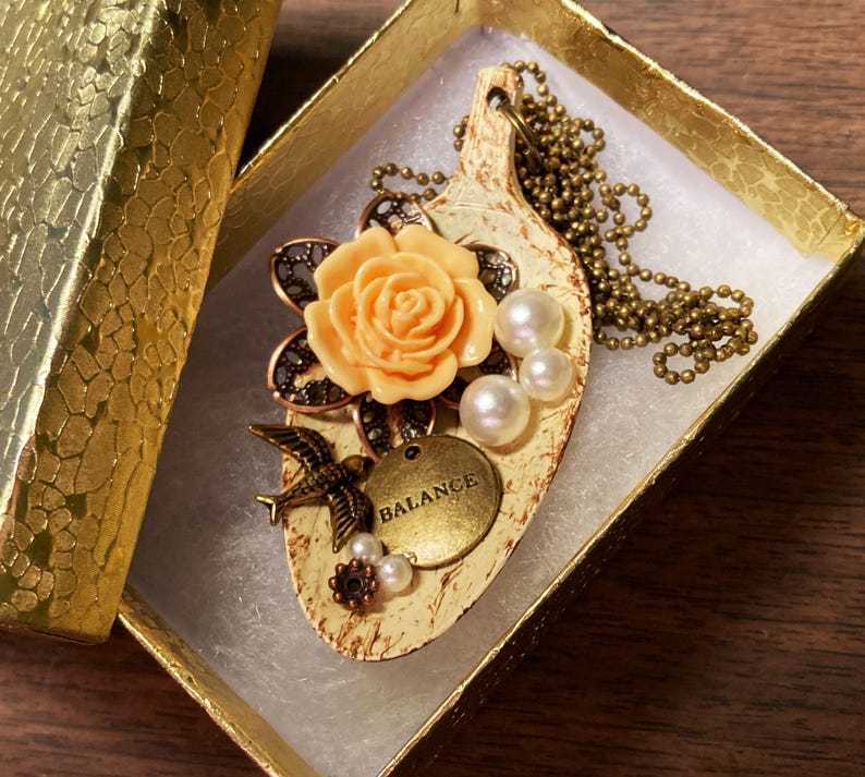 May include: A delicate necklace with a cream-colored pendant featuring a peach-colored rose, a bird, and the word "BALANCE". The pendant is surrounded by pearls and is attached to a gold chain.