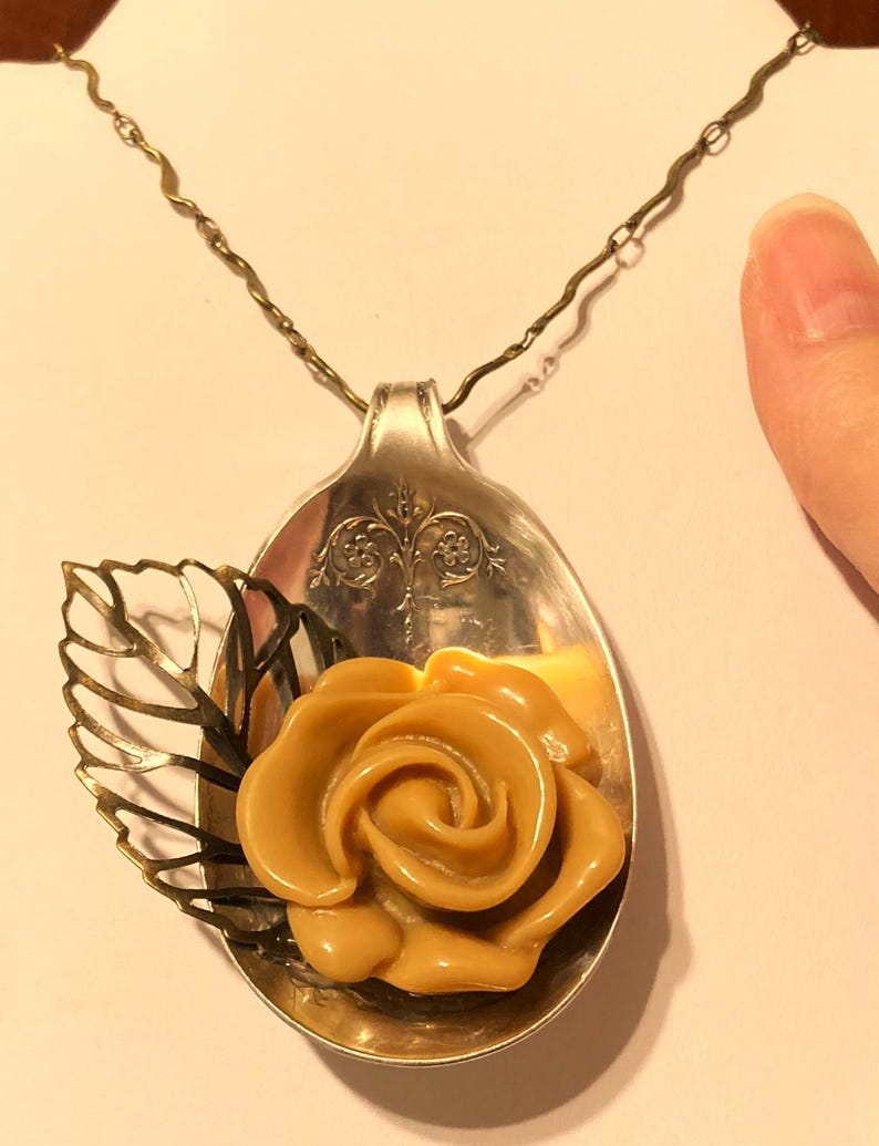 May include: A silver spoon necklace with a gold leaf and a caramel-colored rose pendant.
