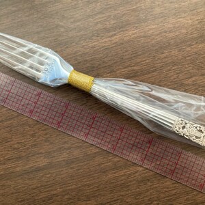 May include: A silver fork with an ornate design on the handle is wrapped in plastic and placed on top of a clear ruler with red markings.