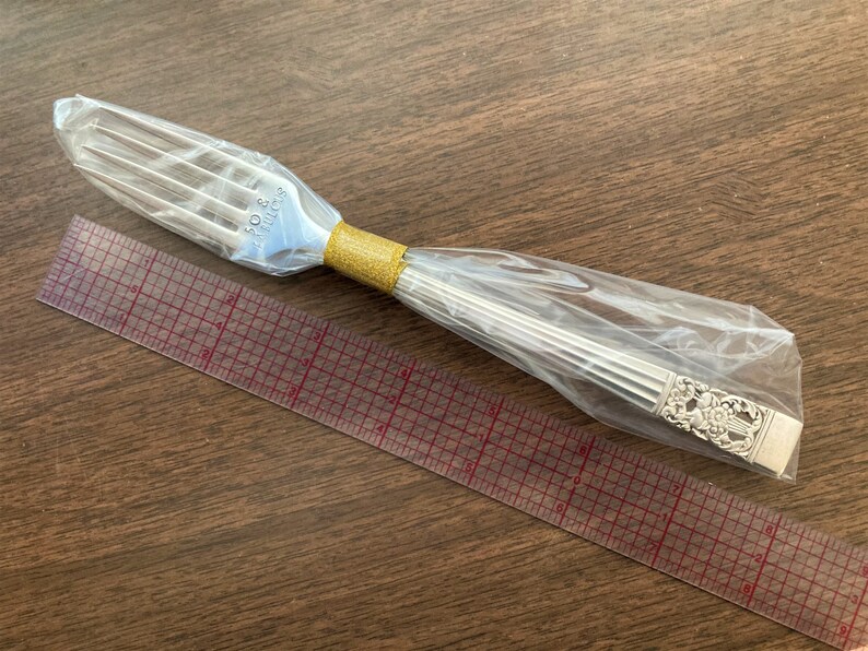 May include: A silver fork with an ornate design on the handle is wrapped in plastic and placed on top of a clear ruler with red markings.