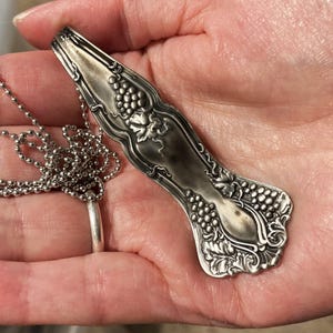 May include: A silver necklace with a pendant shaped like a vintage spoon with grape designs.