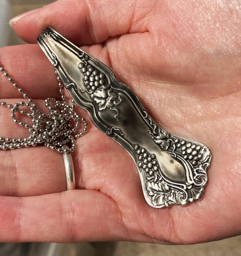 May include: A silver necklace with a pendant shaped like a vintage spoon with grape designs.