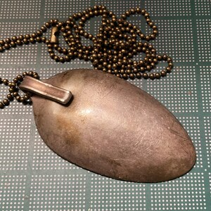 Op de afbeelding: Een zilveren lepelslinger met een messing ketting. De lepel is aan de ketting bevestigd met een metalen ring.