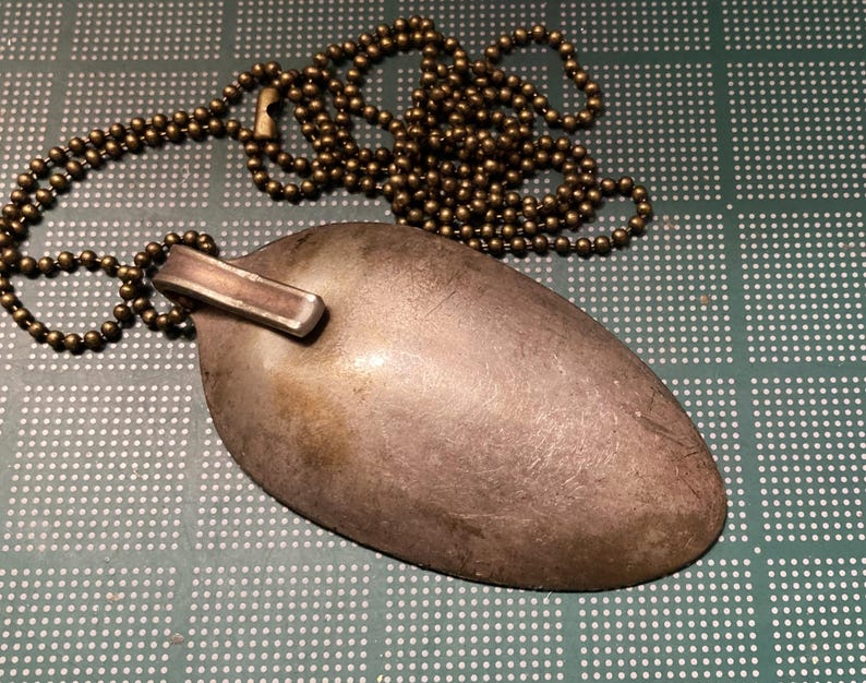 Op de afbeelding: Een zilveren lepelslinger met een messing ketting. De lepel is aan de ketting bevestigd met een metalen ring.