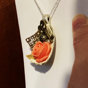 May include: A silver chain necklace with a pendant featuring a peach-colored rose, a key, and a decorative bronze piece. The pendant is shaped like a half-circle.