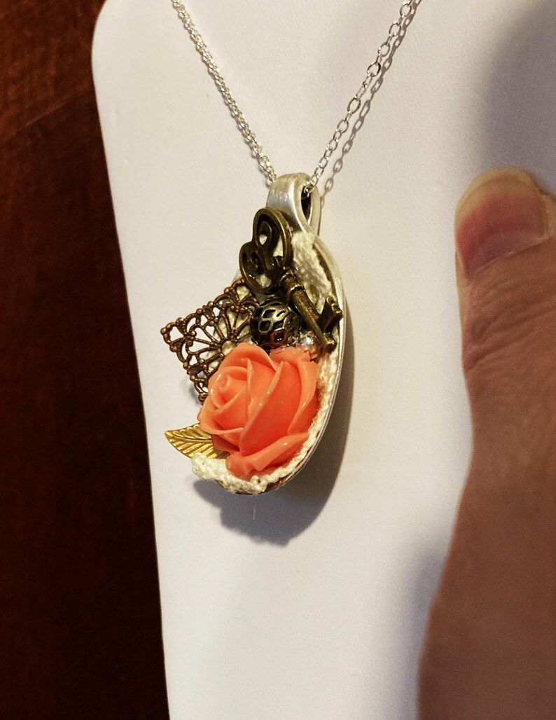 May include: A silver chain necklace with a pendant featuring a peach-colored rose, a key, and a decorative bronze piece. The pendant is shaped like a half-circle.