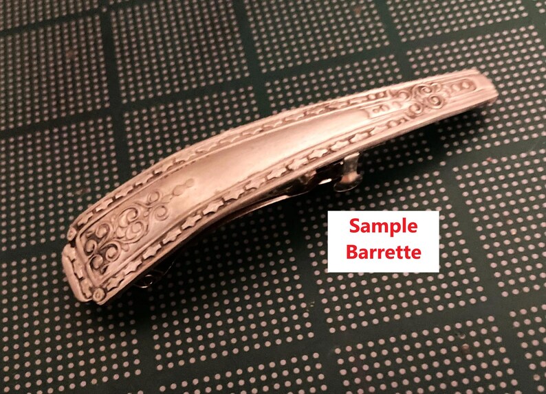 May include: Silver barrette with decorative scrollwork and a hinged clasp. The rectangular barrette has an ornate design along the edges. The text "Sample Barrette" is in a white box.