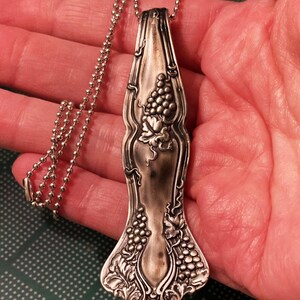 May include: A silver spoon necklace with a grapevine design. The spoon is attached to a silver ball chain.
