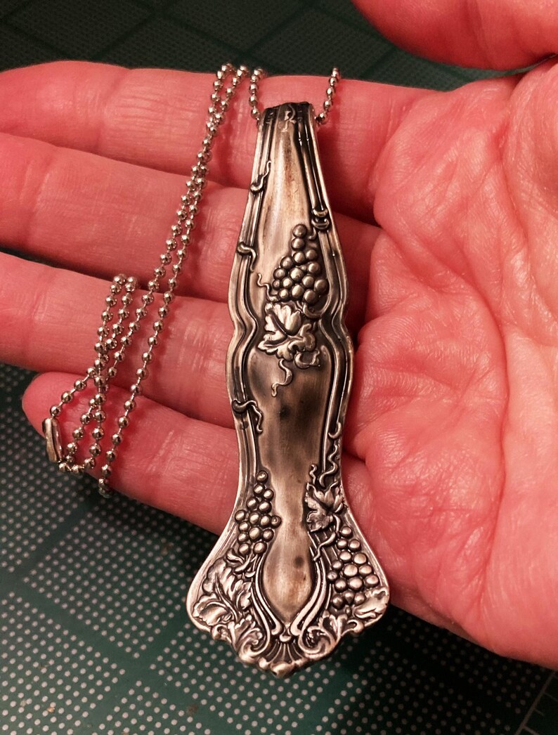 May include: A silver spoon necklace with a grapevine design. The spoon is attached to a silver ball chain.