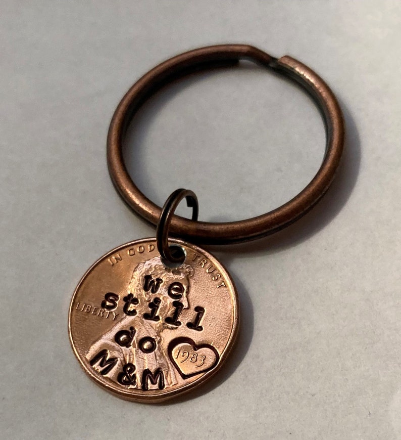 May include: A copper penny keychain with the words "We still do M&M" engraved on it. The penny has a heart with the year 1983 on it.