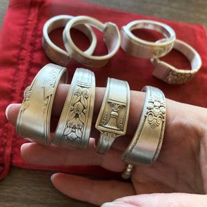 May include: A set of seven silver spoon rings with various decorative designs. The rings are arranged on a red velvet surface and a hand is holding them.
