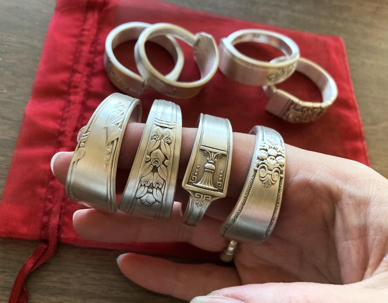 May include: A set of seven silver spoon rings with various decorative designs. The rings are arranged on a red velvet surface and a hand is holding them.