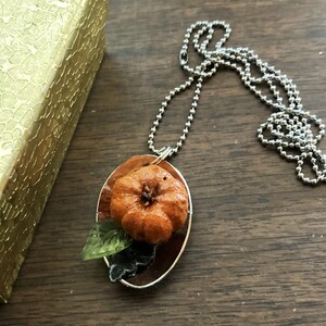 May include: A silver chain necklace with a pendant featuring a small orange pumpkin with green leaves on a silver background.