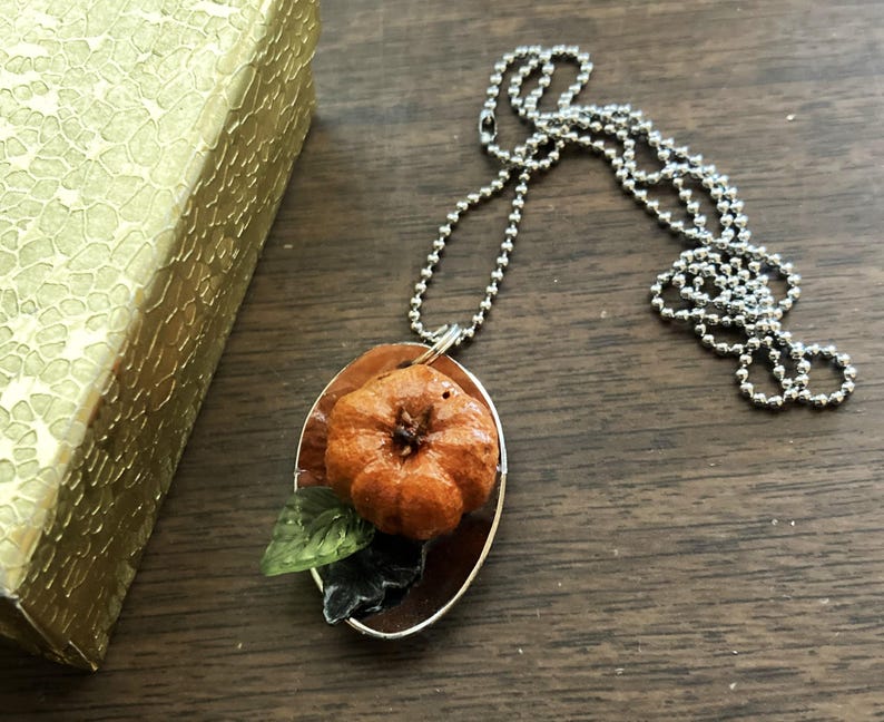 May include: A silver chain necklace with a pendant featuring a small orange pumpkin with green leaves on a silver background.