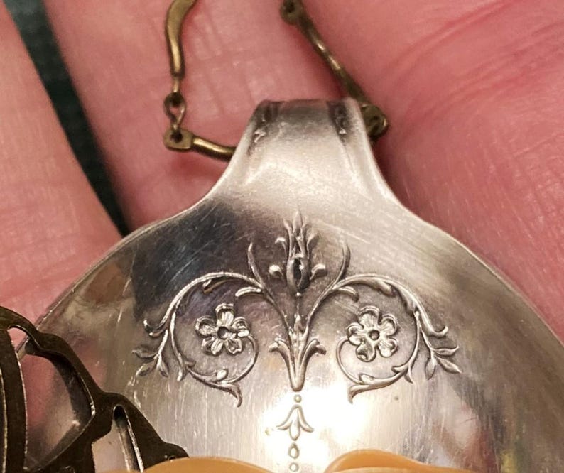 May include: A silver spoon with an ornate floral design, transformed into a pendant with a gold chain.