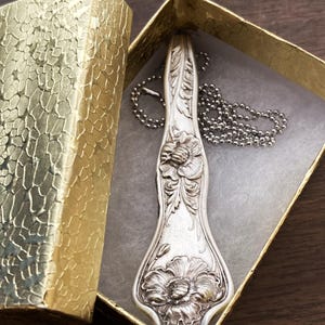May include: A silver spoon necklace with a floral design. The spoon is engraved with two flowers and leaves. The necklace is on a silver chain and is in a small box.