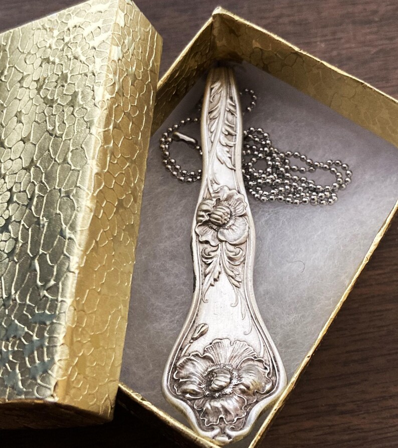 May include: A silver spoon necklace with a floral design. The spoon is engraved with two flowers and leaves. The necklace is on a silver chain and is in a small box.