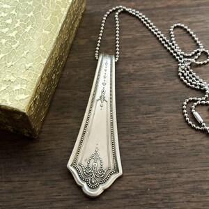 May include: A silver spoon necklace with an ornate design. The spoon is attached to a silver ball chain.