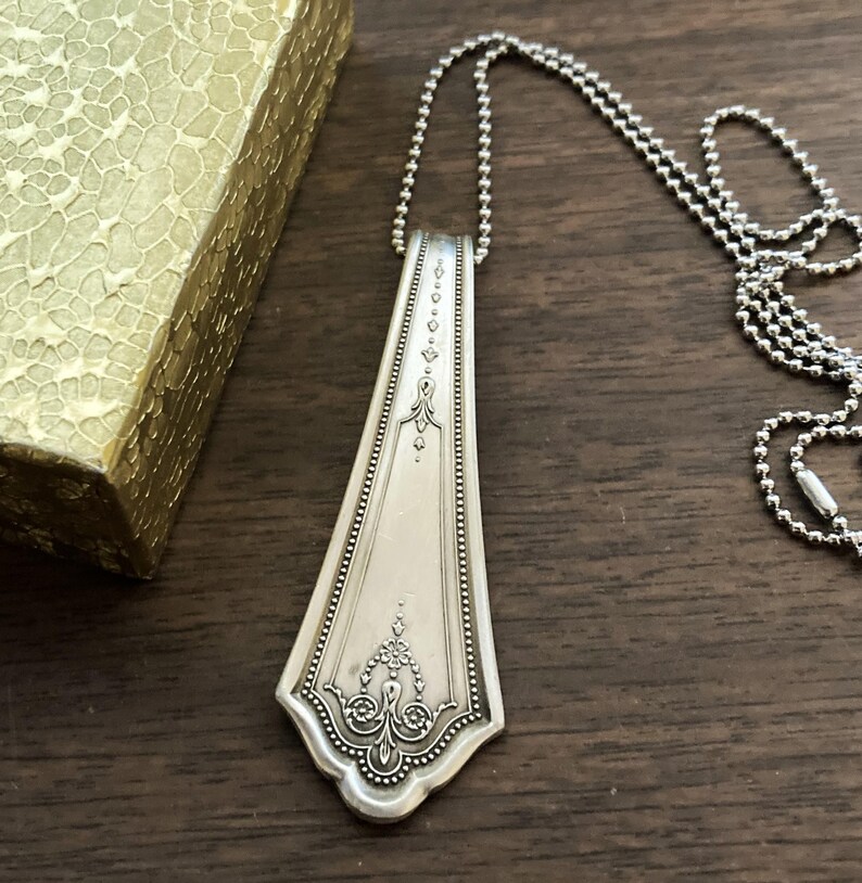 May include: A silver spoon necklace with an ornate design. The spoon is attached to a silver ball chain.
