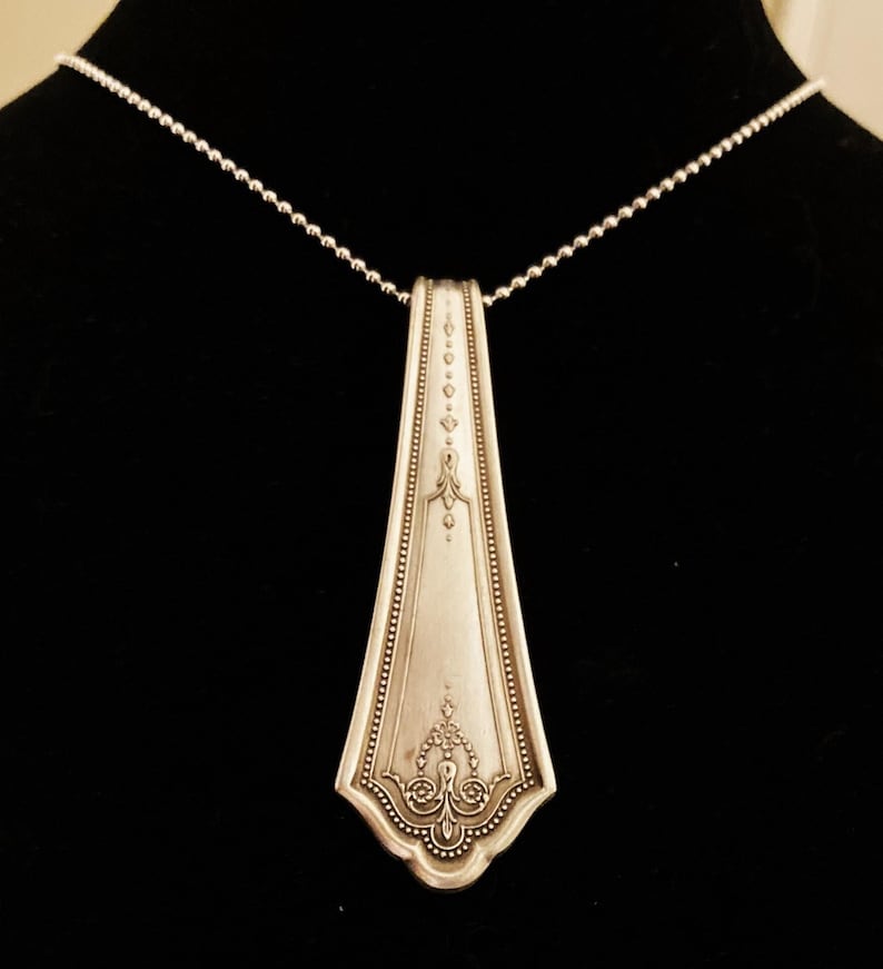May include: A silver necklace with a pendant made from a vintage silverware piece. The pendant is shaped like a long, narrow teardrop with an ornate design.