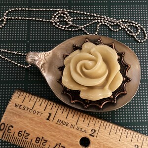 May include: A silver spoon necklace with a cream-colored rose pendant. The rose is surrounded by a copper-colored filigree design.