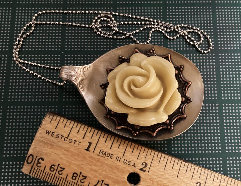 May include: A silver spoon necklace with a cream-colored rose pendant. The rose is surrounded by a copper-colored filigree design.