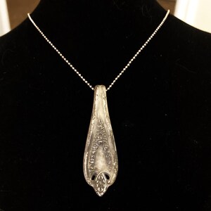 May include: A silver spoon pendant necklace with a delicate design. The pendant is shaped like a teardrop and has a detailed pattern on the front. The necklace is made of a silver chain.