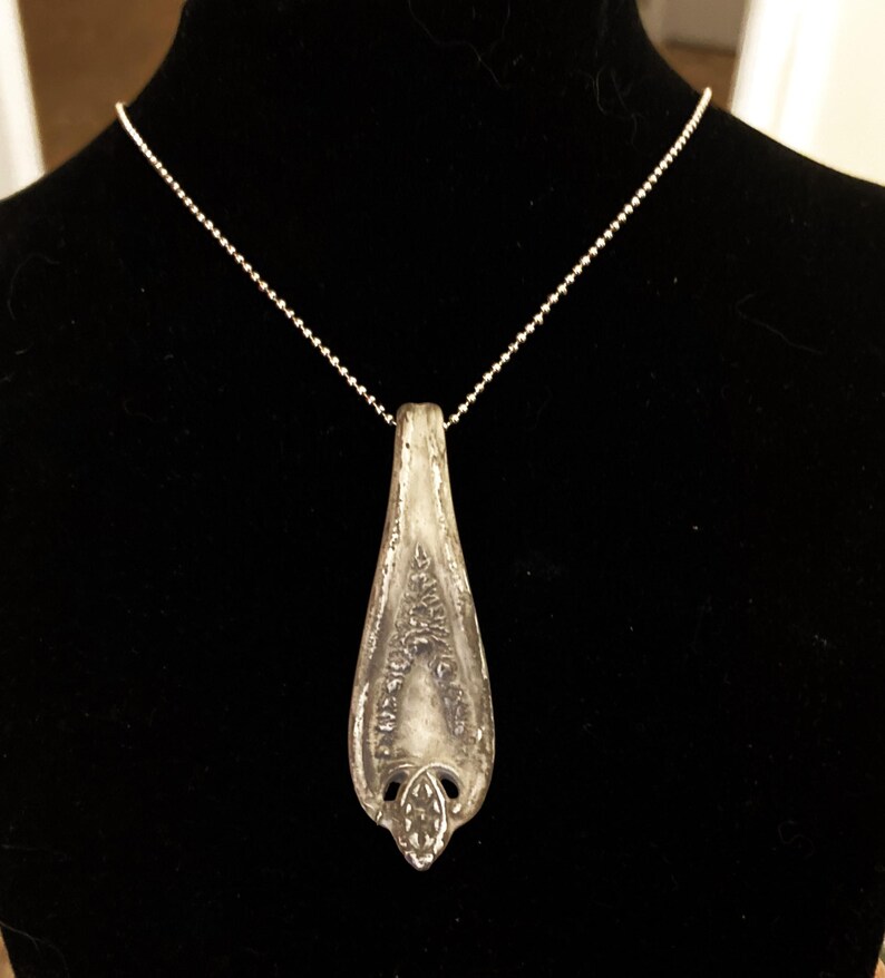 May include: A silver spoon pendant necklace with a delicate design. The pendant is shaped like a teardrop and has a detailed pattern on the front. The necklace is made of a silver chain.