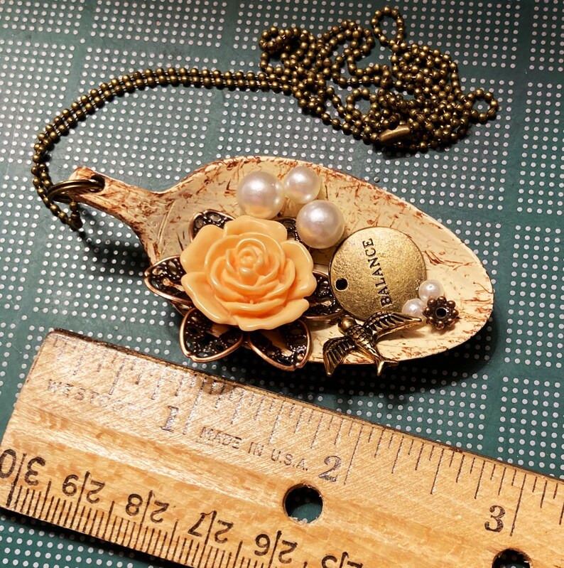 May include: A spoon necklace with a peach-colored rose, white pearls, and a gold charm that says "BALANCE".